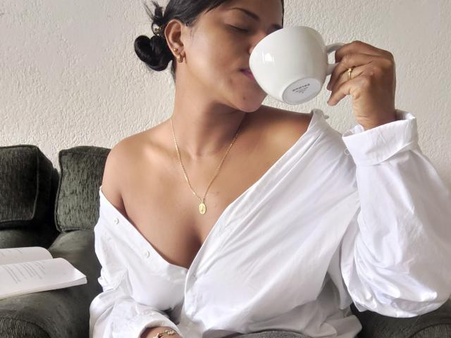 IvannaDuran sips from a white cup, her shirt off-shoulder, exposing her tits, with a gold necklace and ring, sitting on a green couch, looking relaxed.