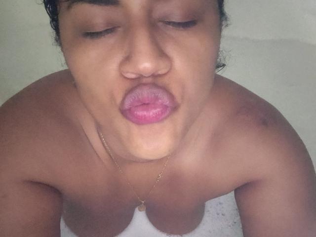 IvannaDuran, topless, with brown skin and pink lips, pouting in a selfie, wearing a gold necklace, eyes closed, small brown blemishes on shoulders, dark curly hair.