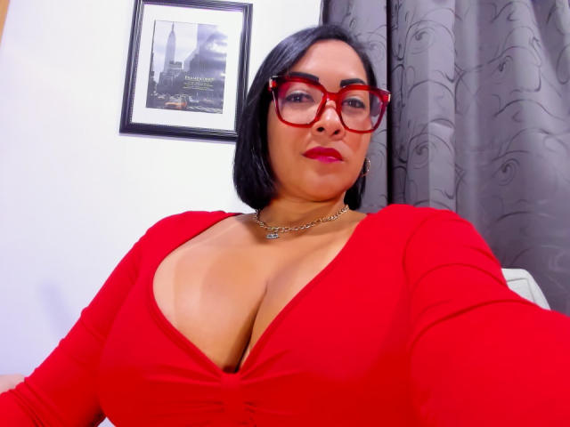 Mellissa Cortez, con gafas rojas, cabello corto negro y una blusa roja que muestra un busto grande, lleva un collar de cadena y está sentada, con una cortina gris de fondo..
