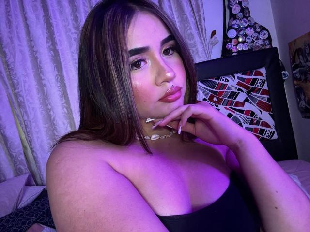 PaulettDelamour has brown hair, pouty pink lips, a nose ring, and is wearing a black strapless top, with her hand near her chin, in purple-lit bedroom.