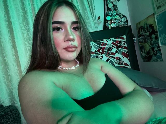 PaulettDelamour with green-tinted skin, long brown hair, thick eyebrows, pink blush, and a white necklace, arms crossed, showing large boobs in a black strap top.