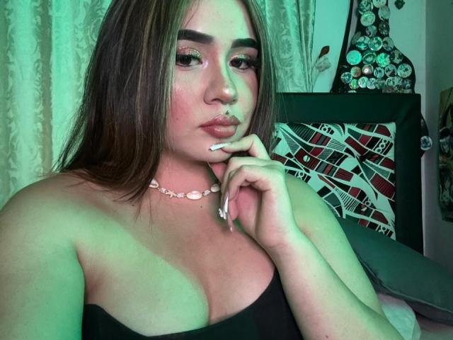 PaulettDelamour with greenish lighting, brown hair, full lips, pink eye makeup, pink necklace, black strap top, showing her tits, hand near mouth, white nails, looking at camera.