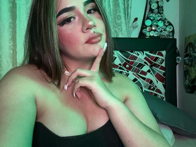 PaulettDelamour with pouty lips, brown hair, green eyeshadow, nose ring, and long nails, touching chin, wearing black strapless top, showing her tits. Bedroom with green lighting.