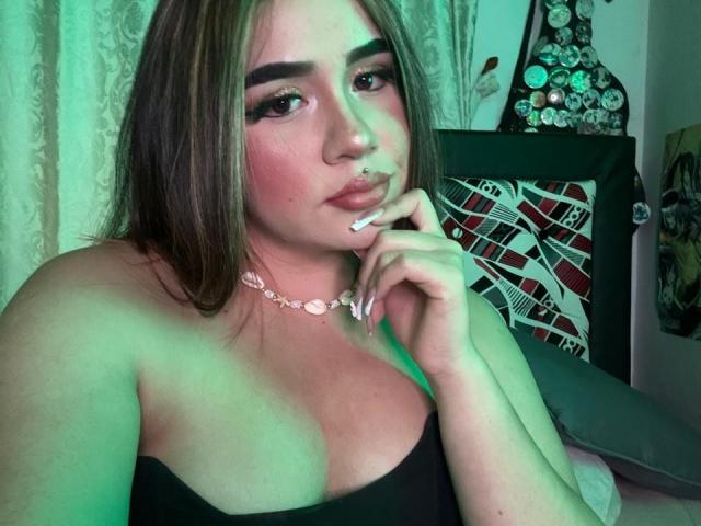PaulettDelamour with green-tinted skin, brown hair, and pink lipstick, holding a cigarette, wearing a black strap top, showing her tits, and a pink necklace.