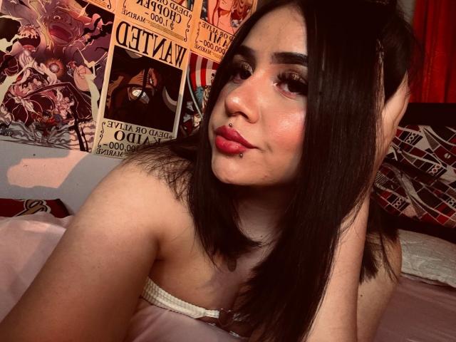 PaulettDelamour with dark hair, pink lipstick, nose piercing, and eyeliner, sitting on bed, wearing strapless pink top, looking at camera with slight smile, showing her tits.