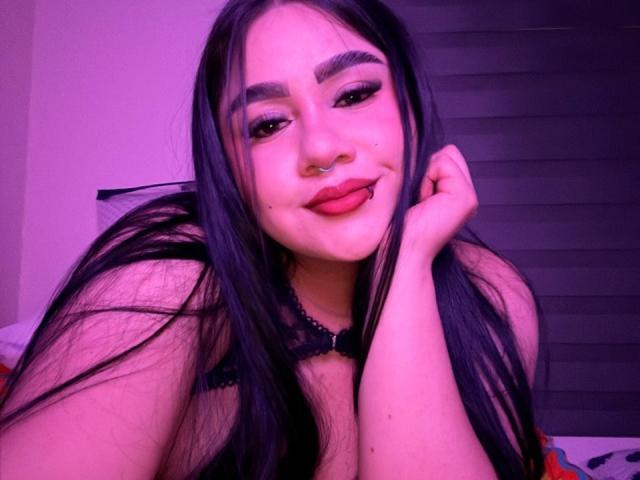 PaulettDelamour with long black hair, nose ring, and red lipstick, smiling with tongue out, hand on cheek, wearing black lace top, in purple-lit room.