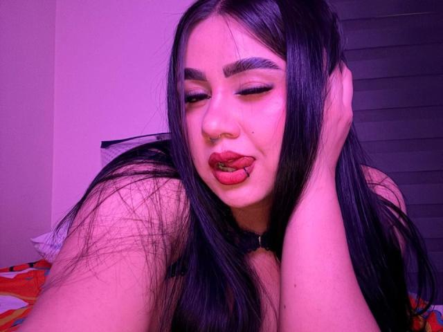 PaulettDelamour with long black hair, pinkish-purple skin, and red lipstick, tongue out, eyes closed, hand in hair, wearing a black choker, in a purple-lit room.