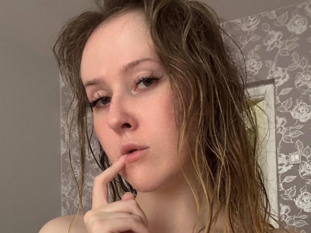 VivanGradney, con el cabello castaño y húmedo, mordiéndose el dedo, tiene labios rosados y un maquillaje sutil, y mira ligeramente hacia abajo, en un selfie frente a una pared gris con un estampado floral..