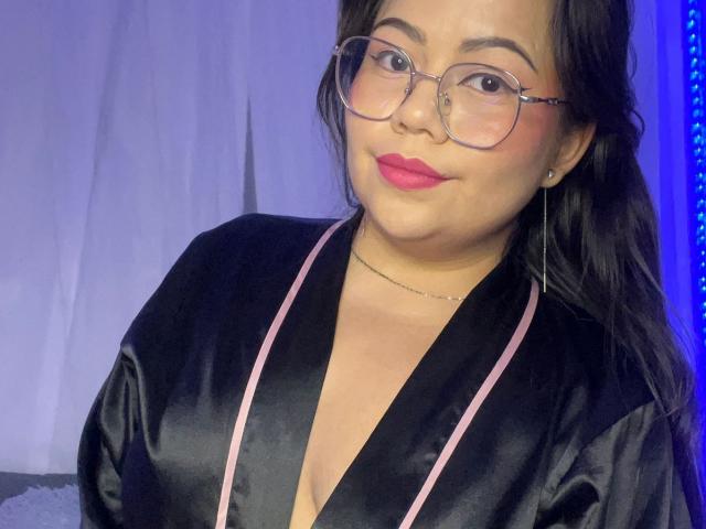 LilyRousee with glasses, black silk robe, pink trim, and pink lipstick, showing a slight smile, her medium tits visible, black hair, and a gold necklace.