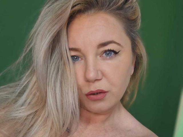 VeraNoires has blonde hair, blue eyes, and pink lips, looking straight at the camera with a slight pout. She's topless, showing her bare shoulders.