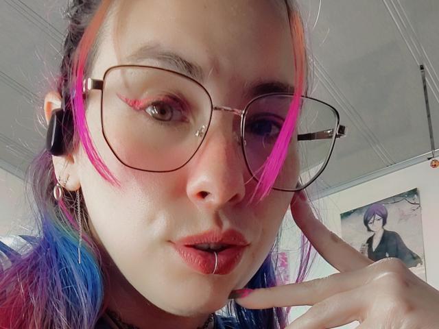 NatashaLau with pink and blue hair, wearing glasses, a nose ring, and a lip ring, pointing at her face with a finger, in a selfie.