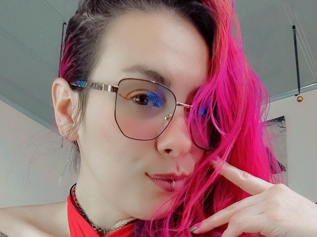 NatashaLau met roze haar, een bril en een rode top, die haar lip aanraakt met een vinger, met een lichte glimlach, en die naar de camera kijkt. Haar borsten zijn klein, ze is schattig..