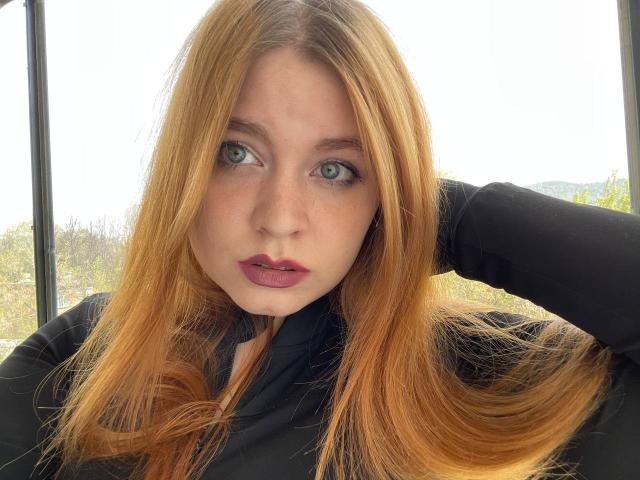 ClemenciaGerhart has long, red-orange hair, blue eyes, and pinkish-red lipstick. She's wearing a black top, with one hand in her hair, looking directly at the camera.