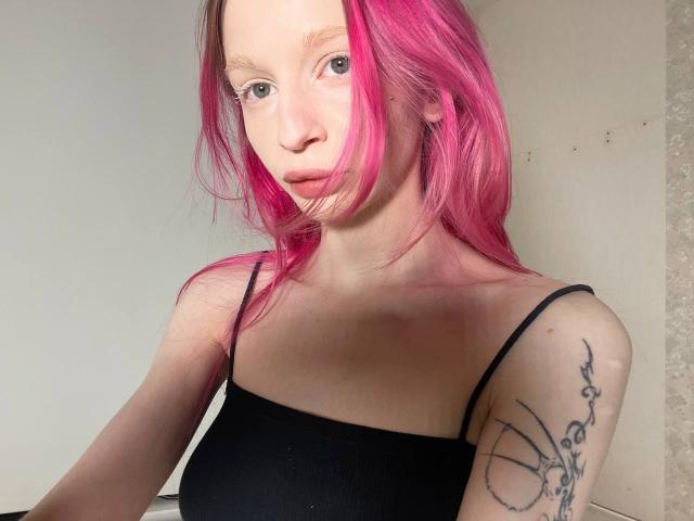 AmiTinn avec des cheveux roses, un haut noir à bretelles spaghetti, un tatouage sur le bras droit, une peau pâle, de petites seins, regardant la caméra, lèvres légèrement entrouvertes, maquillage léger, fond blanc..