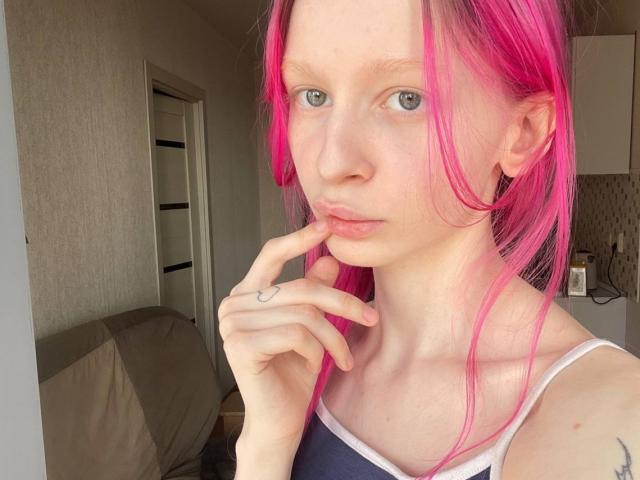 AmiTinn, con cabello rosa, piel pálida y ojos azules, tocándose los labios con un dedo, vistiendo una blusa azul y blanca, mostrando un pequeño tatuaje en su dedo..