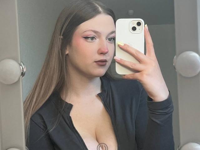 NadaHenig, de piel pálida, cabello castaño y maquillaje, sosteniendo un teléfono, vistiendo una blusa negra, mostrando sus senos, uñas amarillas y un piercing en la nariz, en un selfie frente al espejo..