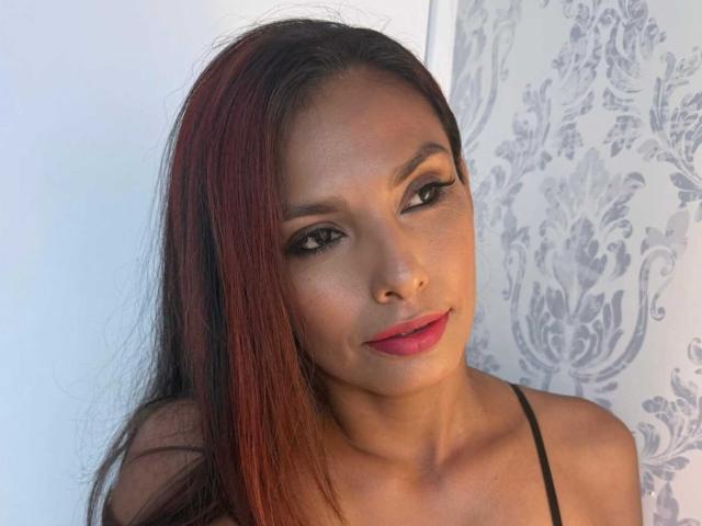 RoxaneMoore with dark brown, red-highlighted hair, wearing black spaghetti straps, has full lips with pink lipstick, and subtle makeup, smiling slightly at the camera.