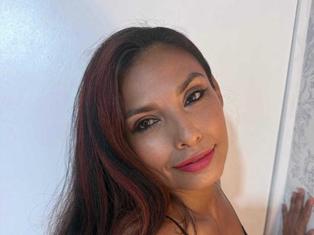 RoxaneMoore hat dunkelbraunes Haar mit roten Strähnen, schwarzen Eyeliner und pinkem Lippenstift, lächelt in die Kamera und neigt leicht den Kopf, wobei sie ihr süßes Gesicht und ihre Brüste zeigt..
