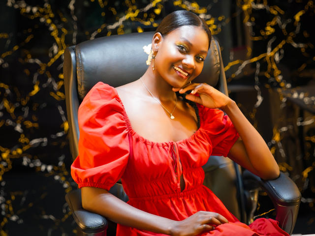 Andrea, une femme à la peau foncée, avec de petites seins, une robe rouge, des manches bouffantes, souriante, une main sur la joue, assise sur un fauteuil en cuir noir, avec des boucles d'oreilles dorées, un collier, et des cheveux noirs, sur un fond sombre et à motifs..