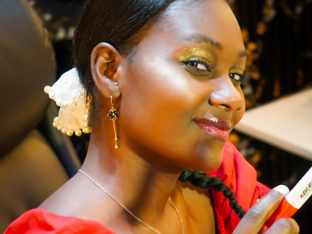 AndreaStorm avec un maquillage des yeux doré brillant, une robe rouge, des tresses noires, un accessoire pour les cheveux blanc, des boucles d'oreilles pendantes, tenant un pinceau de maquillage, souriant avec des lèvres roses brillantes..