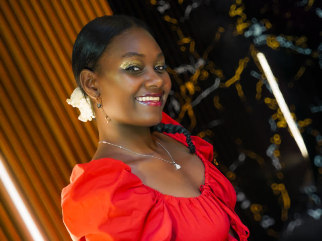 AndreaStorm with dark skin, red puffy-sleeve dress, braided hair, white flower, earrings, necklace, smiling, bright green eye makeup, shiny red lips, medium-sized tits, warm lighting.