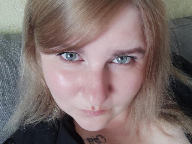 MinistraX has pale skin, blue eyes, and blonde hair with side bangs. She's pouting slightly, with a small tattoo visible below her chin. Her face is shiny and she's wearing black.