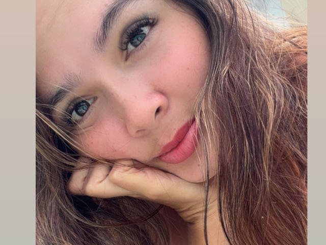 Gabrielafoxxy's face with blue eyes, pink lips, and brown wavy hair, resting her chin on her hand, looking slightly to the side. Her skin is fair with a few freckles.