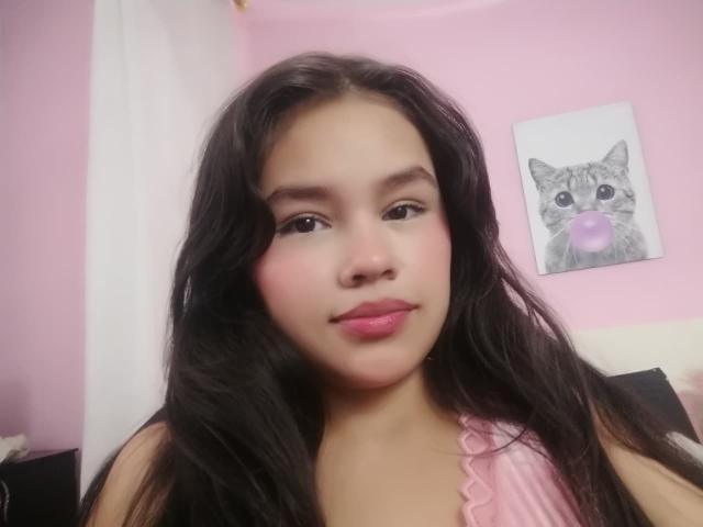 RubyCrow with long black hair, pink lips, and a slight smile, wearing a pink top, has a cute cat painting in the pink-walled background, blowing pink bubble gum.
