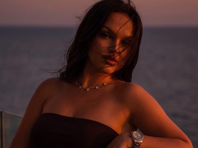 JaneOSweetAh with dark hair, wearing a strapless black top, necklace, and watch, has a sexy, confident look, ocean in the background, sunset lighting, slightly windswept face.