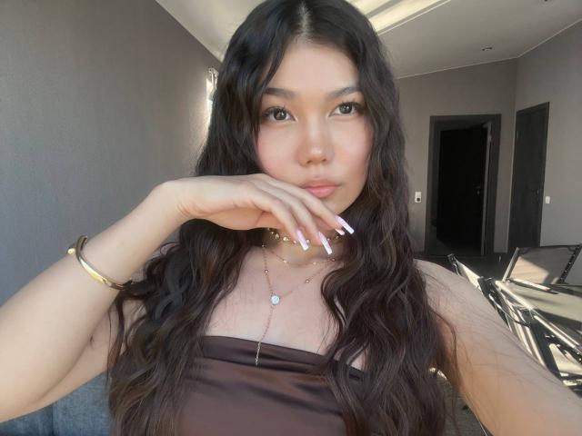 Ameliya Ayrton, com longos cabelos escuros e ondulados, um top marrom sem alças, uma pulseira dourada e unhas longas brancas, com a mão apoiada no rosto, olhando para a câmera..