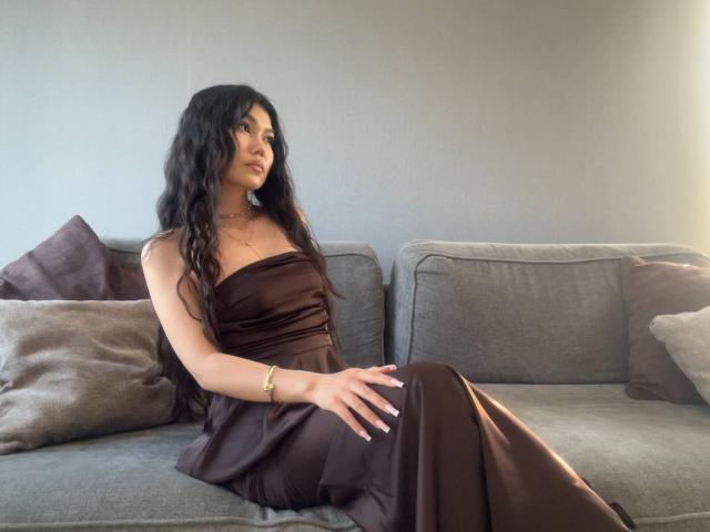 Ameliya Ayrton, com longos cabelos pretos ondulados, vestindo um vestido marrom de cetim sem alças, sentada em um sofá cinza, com a mão na coxa, olhando para o lado, com luz vindo da direita..