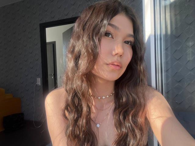Ameliya Ayrton hat welliges, braunes Haar, eine niedliche Nase und zieht die Lippen leicht zusammen. Sie trägt eine Halskette und ist obenhin nackt, wobei ihre kleinen Brüste zu sehen sind..