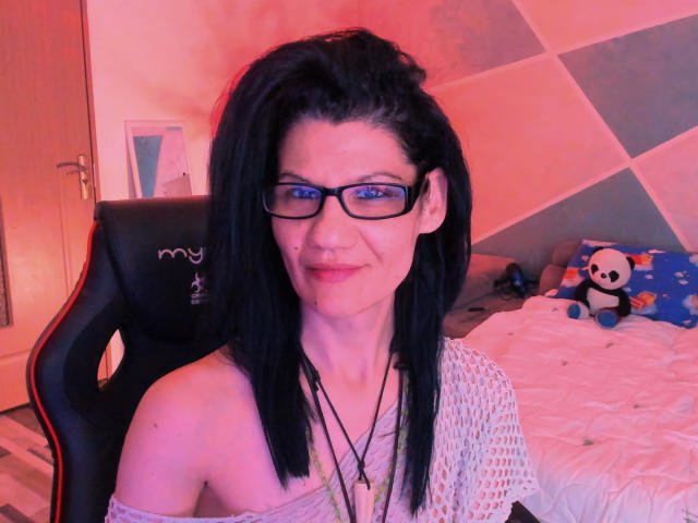 Vanessa69Xo, com cabelo preto, óculos e uma blusa de malha branca, sentada em uma cadeira preta, com um pequeno ponto no lábio superior e um colar. Cenário de quarto..