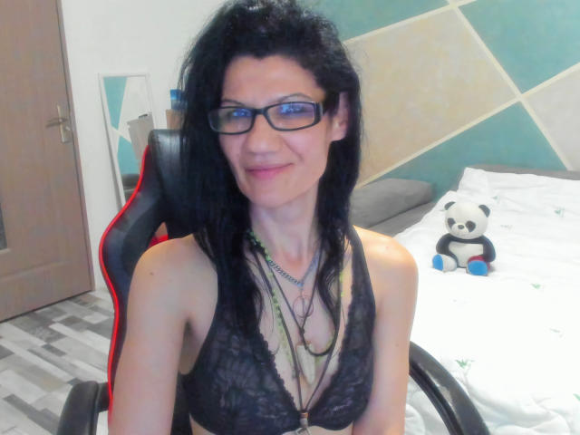 Vanessa69Xo con reggiseno nero in pizzo, occhiali e lunghi capelli neri, seduta su una sedia rossa e nera, sorridente, che indossa una collana e ha un piccolo giocattolo a forma di panda sul letto..