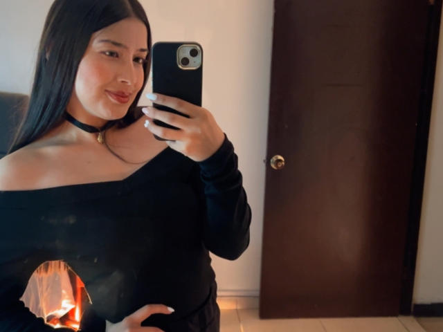 EmmaLombardi with long black hair, black off-shoulder top, black choker, holding a lit candle, taking a mirror selfie, smiling, wearing dark pants.