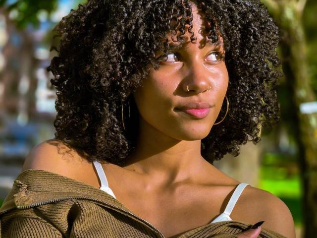 CinthiaWalker a les cheveux noirs bouclés, un anneau de nez et des boucles d'oreilles. Elle porte un haut vert à une épaule, laissant apparaître les bretelles blanches de son soutien-gorge, et regarde sur le côté avec un léger sourire..