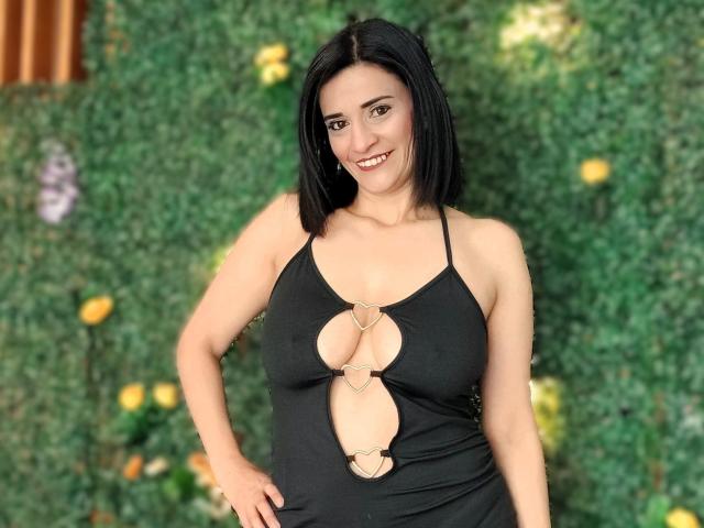 MaraRoberttss, con un corte de pelo bob negro, sonriendo, vestida con un vestido negro de tirantes con diseños de corazones recortados sobre sus grandes senos. Fondo verde, con las manos en las caderas..