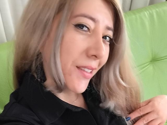 HotPamelaSex has blonde hair, black eyeliner, and blue nails, smiling with pink lips, wearing a black shirt, sitting on a green couch.