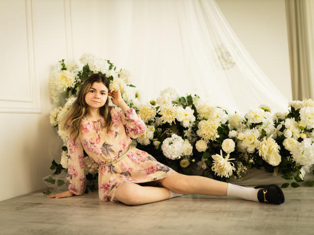TinaRedly, seduta con le gambe divaricate, vestito floreale, capelli lunghi, viso carino, seno piccolo, calze bianche, scarpe nere, appoggiata a un muro di fiori, con una mano tra i capelli, sorridente..