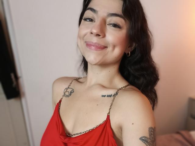 AnntonellaWatson with wavy black hair, smiling, red dress, small tits, tattoos on chest and arm, gold hoop earrings, light skin, pink lipstick, in a well-lit room.