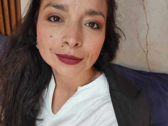 StangelarRivera has dark wavy hair, brown eyes, and red lipstick. She's wearing a white shirt with a grey jacket. Her face has a few moles and a small nose piercing.