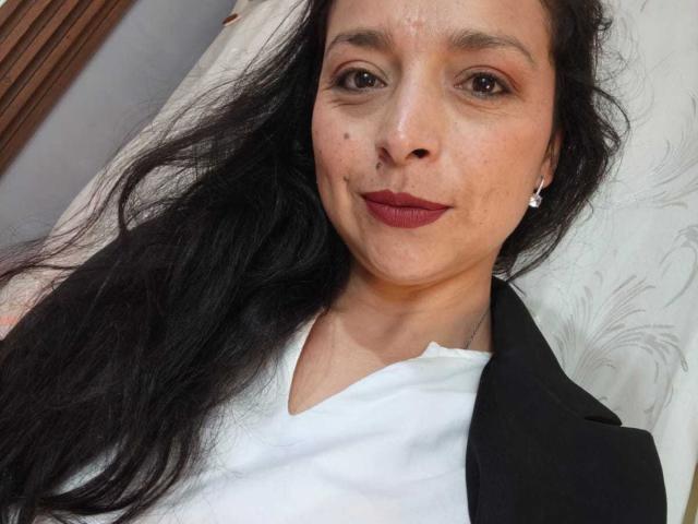 StangelarRivera, con cabello negro largo, ojos marrones y lápiz labial rojo, vestida con una camisa blanca y una chaqueta negra, acostada, mostrando una sonrisa sutil, con pequeños aretes de diamantes..