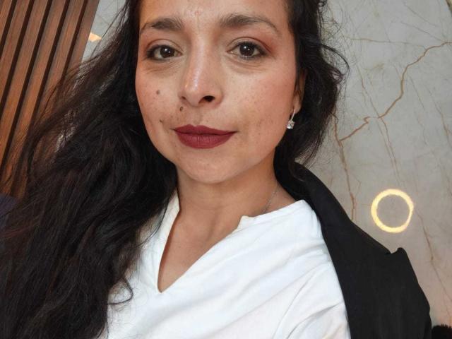 StangelarRivera ha la pelle marrone media, lunghi capelli neri, rossetto rosso, piccole lentiggini sul viso e indossa una camicia bianca con una giacca nera e orecchini di diamanti..