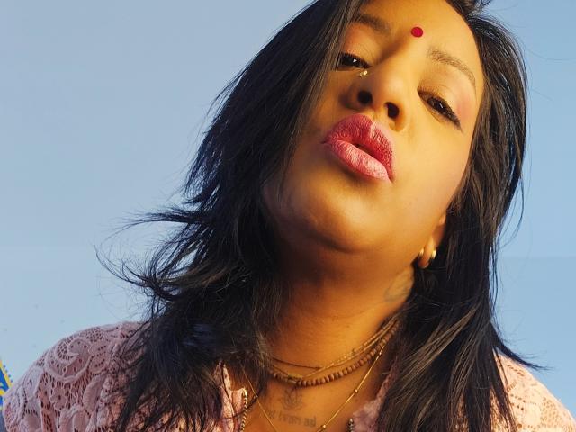 Nahirr has medium brown skin, black wavy hair, red bindi on forehead, gold hoop earrings, pink lace shirt, multiple gold necklaces, and pink glossy lips, looking slightly down.