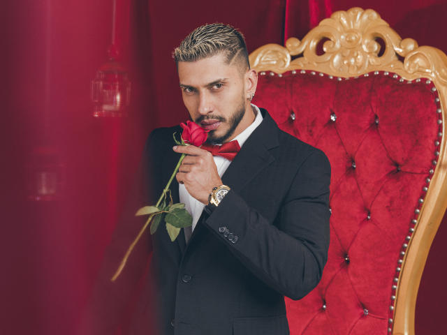SethVegas with short blonde hair, holding a red rose to his nose, wearing a black suit, red bowtie, white shirt, and gold watch, standing in front of a red velvet chair.