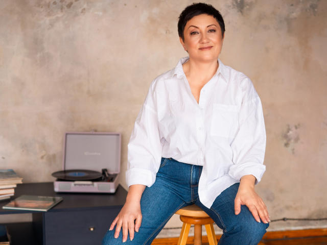 EmilyLlove with short dark hair, wearing a white button-up shirt and blue jeans, sitting on a wooden stool, hands on her thighs, smiling softly.