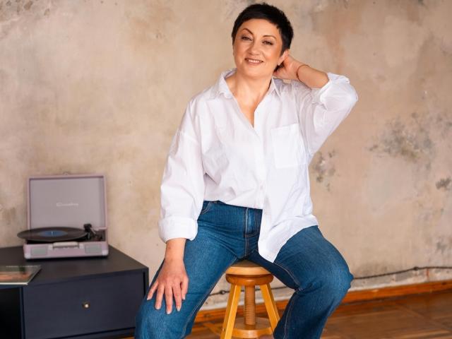 EmilyLlove with short brown hair, white button-up shirt, and blue jeans, sitting on a wooden stool, smiling, hand in hair, in a casual, relaxed pose.
