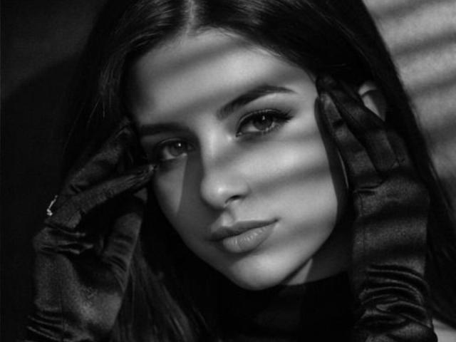 Black-and-white photo of IsabellaJames with long dark hair, wearing glossy black gloves, touching her face, light shadows across her face, looking seductively at the camera.