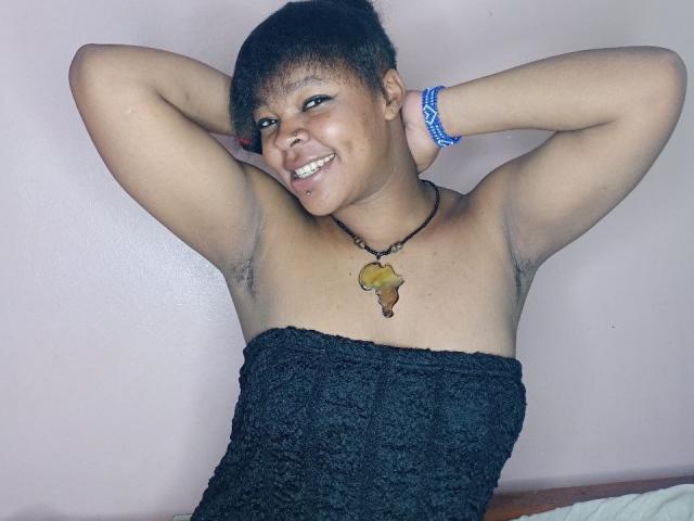 RareGem with short black hair, smiling, arms raised, showing armpit hair, wearing a black textured tube top, necklace with yellow pendant, blue bracelet, in a pink-walled room.
