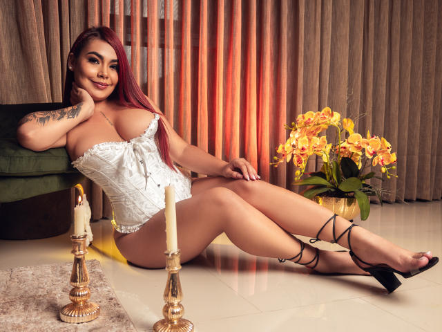 ValeryJhonston, avec des cheveux roux, un corset en dentelle blanche, des talons hauts noirs à brides, un tatouage sur le bras, assise par terre, les jambes croisées, des orchidées jaunes, des bougies, un canapé vert, un sol carrelé brillant, souriante..